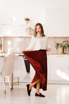 Portrait of fashionable woman in a red skirt, white blouse and stylish suede shoes with a buckle posing on the kitchen. Girl with a big red lips