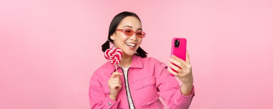 Portrait of stylish, happy asian girl taking selfie with candy, lolipop sweets and smiling, taking photo with mobile app, standing over pink background.