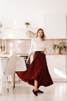 Portrait of fashionable woman in a red skirt, white blouse and stylish suede shoes with a buckle posing on the kitchen. Girl with a big red lips