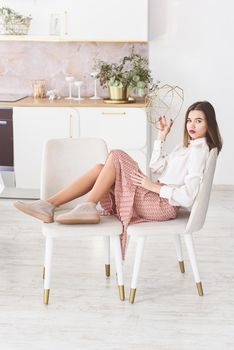 Portrait of fashionable women in beige skirt, white blouse and stylish suede loafer posing on the kitchen in a chair. Girl with a big red lips