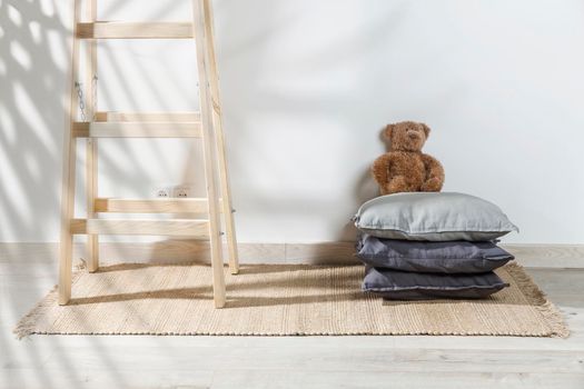 The teddy bear is leaning against the wall next to a small children's stool with three gray pillows on it. The stepladder is leaning against the wall. Copy space