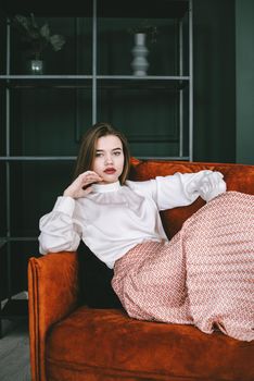 Portrait of fashionable young woman in a beige skirt and white blouse posing on a orange sofa. Girl with a big dark red lips