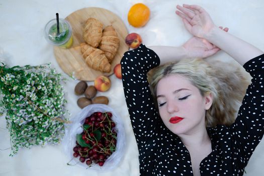 charming blonde young woman on a picnic on plaid in park with tasty snacks. Lemonade, fruits and croissants. summertime, rest, relax, enjoy. freedom.