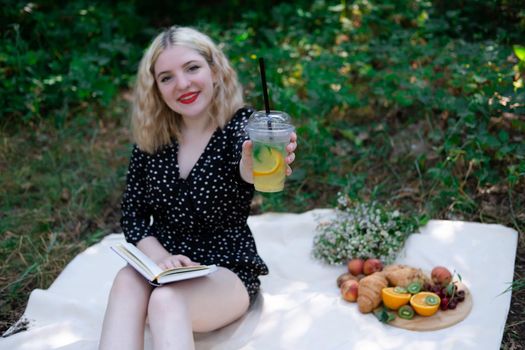 portrait of young woman on a picnic on plaid in park reading a book with tasty snacks.