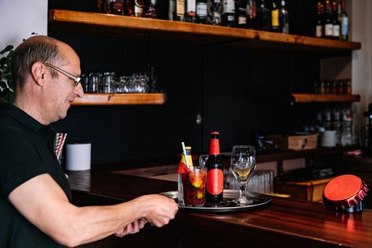 Male waiter, mature, bald, smiling, dressed in company uniform, black polo shirt, Mature, experienced waiter taking an order at the bar counter. worker in his small busines. Worker in his small business. Warm atmosphere and dim lighting. Horizontal