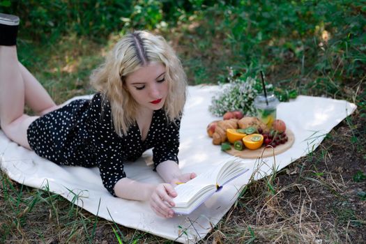 portrait of young woman on a picnic on plaid in park reading a book with tasty snacks.