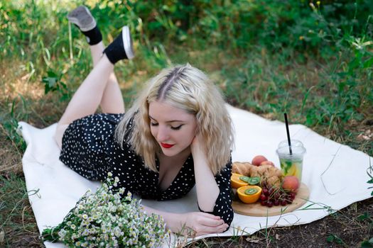 charming blonde young woman on a picnic on plaid in park with tasty snacks. Lemonade, fruits and croissants. summertime, rest, relax, enjoy. freedom.