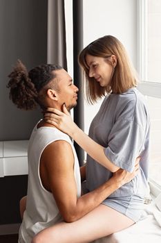 A multiracial couple, a guy and a girl, in cozy home clothes, sit face to face, near the window