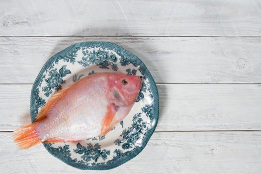 raw pink tilapia fish lies on a plate with blue ornaments on white wooden boards, copy space. High quality photo