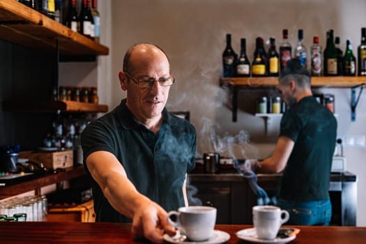 Two co-workers in their small business. Mature waiter, experienced and smiling, bald, with black polo shirt and glasses. Young waiter with long hair tied back and smiling, wearing black polo shirt. Smiling partners of a small business standing, preparing coffee for their customers. Warm and pleasant atmosphere, natural light. Horizontal