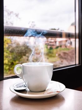 Close up shot of a white coffee cup on a bar table with a window in the background. Cup of steaming coffee. Warm and pleasant atmosphere, natural light, cloudy day. Vertical