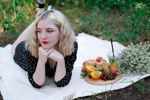 pretty blonde girl on a picnic in a forest or park with lemonade, fruits and croissants. summertime, rest, relaxation. millennial generation.