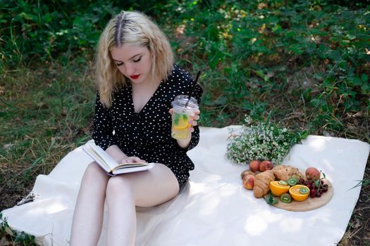 portrait of young woman on a picnic on plaid in park reading a book with tasty snacks.