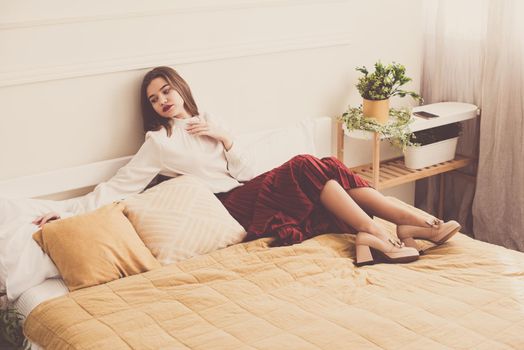 Portrait of fashionable women in red skirt, white blouse and stylish beige high-heeled shoes with a chain buckle posing in a bed. Girl with a big red lips