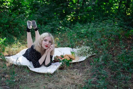 charming blonde young woman on a picnic on plaid in park with tasty snacks. Lemonade, fruits and croissants. summertime, rest, relax, enjoy. freedom.