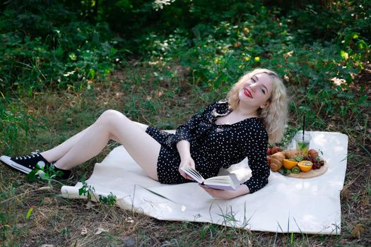 portrait of young woman on a picnic on plaid in park reading a book with tasty snacks.