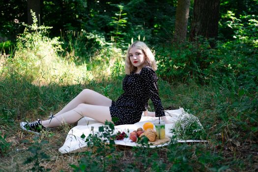 charming blonde young woman on a picnic on plaid in park with tasty snacks. Lemonade, fruits and croissants. summertime, rest, relax, enjoy. freedom.