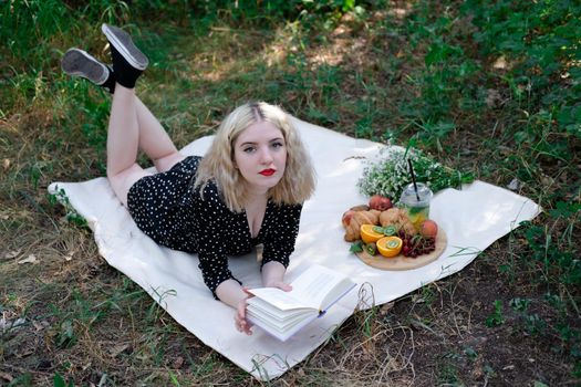 portrait of young woman on a picnic on plaid in park reading a book with tasty snacks.