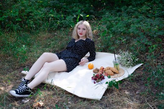 pretty blonde girl on a picnic in a forest or park with lemonade, fruits and croissants. summertime, rest, relaxation. millennial generation.