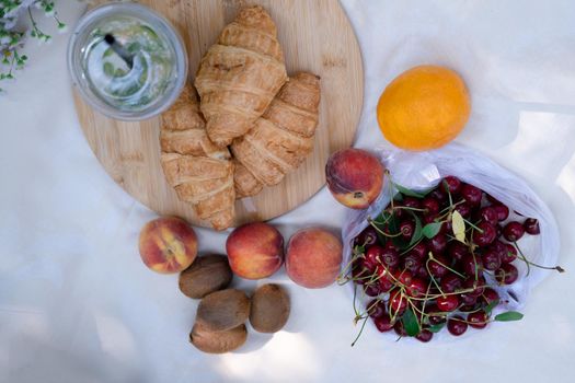 Healthy picnic for a summer day with croissants, fresh fruits and lemonade. peach, cherry, kiwi, oranges. white wildflowers.