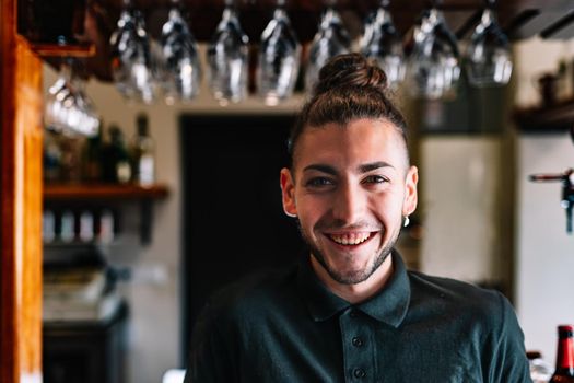 Portrait of a young, smiling, experienced, focused and hard-working waiter, dressed in company uniform, black polo shirt, standing in his small business. Looking at the camera. Warm atmosphere and dim lighting. horizontal Glasses hanging in the background over the counter.