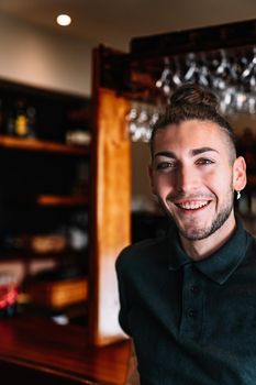 Portrait of a young, smiling, experienced, focused and hard-working waiter, dressed in company uniform, black polo shirt, standing in his small business. Looking at the camera. Warm atmosphere and dim lighting. Vertical. Glasses hanging in the background over the counter.