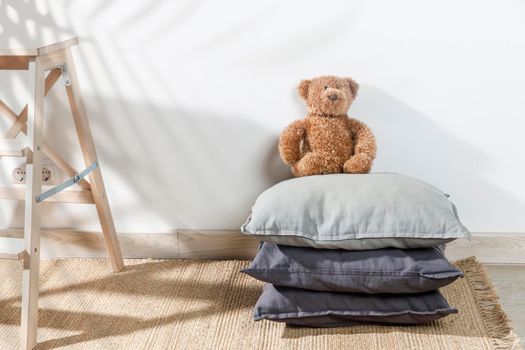 The teddy bear is leaning against the wall next to a small children's stool with three gray pillows on it. The stepladder is leaning against the wall. Copy space