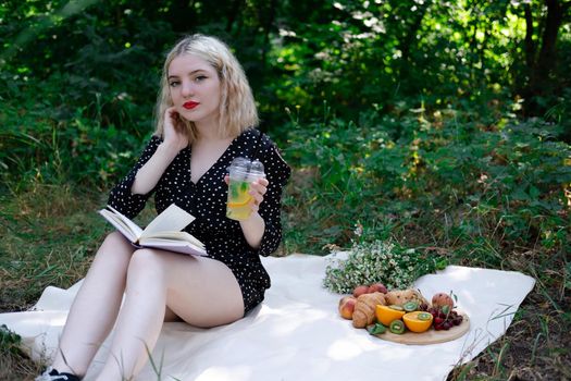 portrait of young woman on a picnic on plaid in park reading a book with tasty snacks.