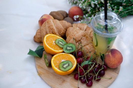 Healthy picnic for a summer day with croissants, fresh fruits and lemonade. peach, cherry, kiwi, oranges. white wildflowers.