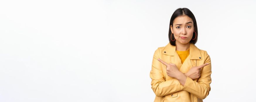 Image of indecisive asian girl, pointing fingers sideways, pointing left and right, choosing variant, deciding, standing over white background. Copy space