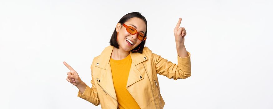 Smiling asian brunette woman in sunglasses, pointing fingers sideways, left and right, showing variants, laughing and dancing, wearing sunglasses, white background.