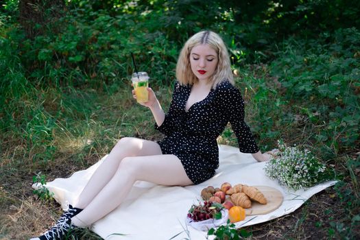 charming blonde young woman on a picnic on plaid in park with tasty snacks. Lemonade, fruits and croissants. summertime, rest, relax, enjoy. freedom.