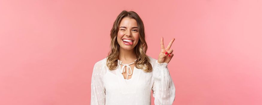 Close-up portrait of friendly joyful blond girl in white dress, sending positive vibes, smiling pleased and wink, stick tongue showing peace sign, looking kawaii over pink background.