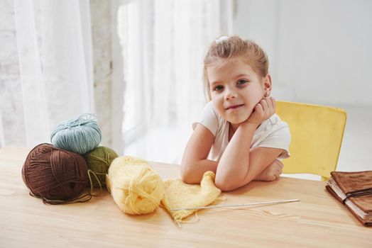 So many threads. Kid is knitting at home. Cute little girl sitting near the wooden table is learning some new stuff.