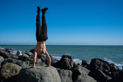 Shirtless man doing handstand on rocks by the sea