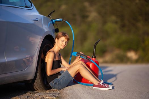 Sad girl sitting on the ground next to electric car. Holding charging cable and red gassoline canister.