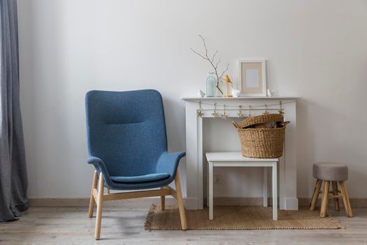 Fragment of the interior of a cozy house. A gray armchair by a white fireplace. On the fireplace is a glass bottle with a branch, figurines of birds and a photo frame. Place for text