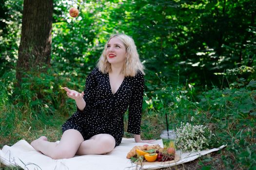 pretty blonde girl on a picnic in a forest or park with lemonade, fruits and croissants. summertime, rest, relaxation. millennial generation.