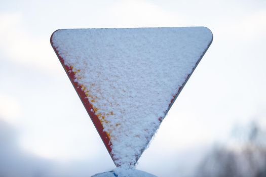 Road sign covered with a layer of white and cold snow. Give way to priority. Against the background of a blurred sky. The shape of a triangle. Road signs warning.