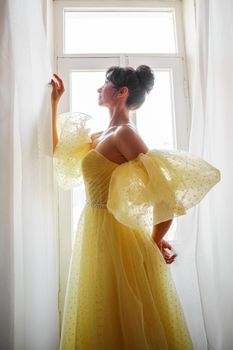 A woman's silhouette in a golden luxurious dress against the background of a window holds a curtain with her hands. Elegant lady in a yellow long silk dress with bare back, back view