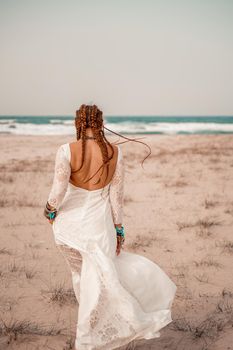 Model in boho style in a white long dress and silver jewelry on the beach. Her hair is braided, and there are many bracelets on her arms