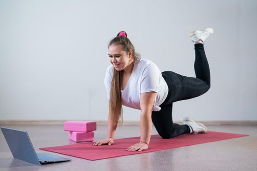 Young woman doing sports exercises at home. The girl is engaged in fitness at home on the mat and watches a training video on a laptop.