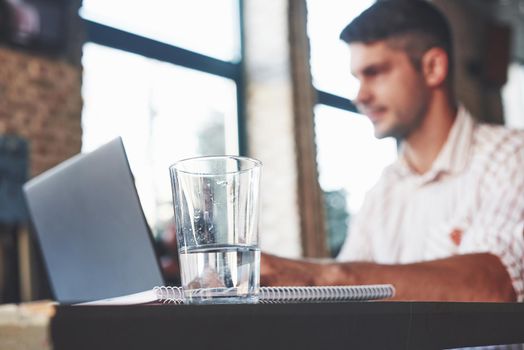 Glass of water in focus. Adult man sits in cafe at daytime and using the laptop for the remote work.