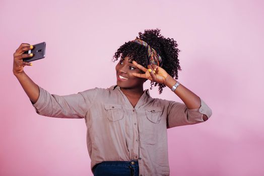 Photo of the day. Attractive afro american woman in casual clothes takes selfie and shows gesture with two fingers at pink background in the studio.