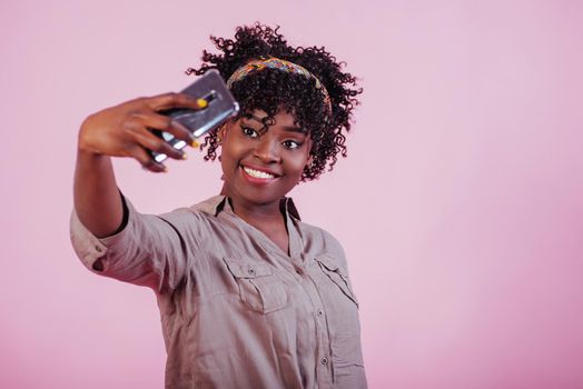 Playful mood. Attractive afro american woman in casual clothes takes selfie at pink background in the studio.