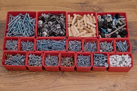 Various size and shape of screws, bolts, wooden pegs selection in red plastic tray box on a wooden background. Assembly kit for furniture.