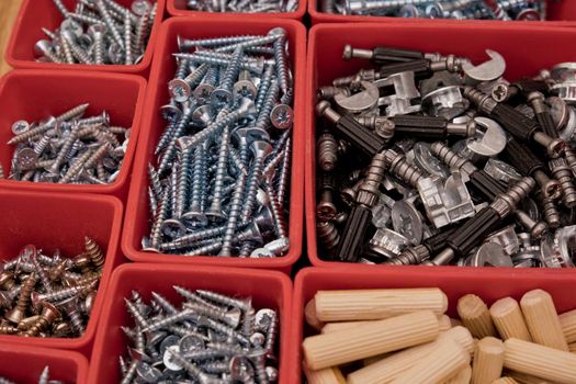 Various size and shape of screws, bolts, wooden pegs selection in red plastic tray box on a wooden background. Assembly kit for furniture. Selective focus.