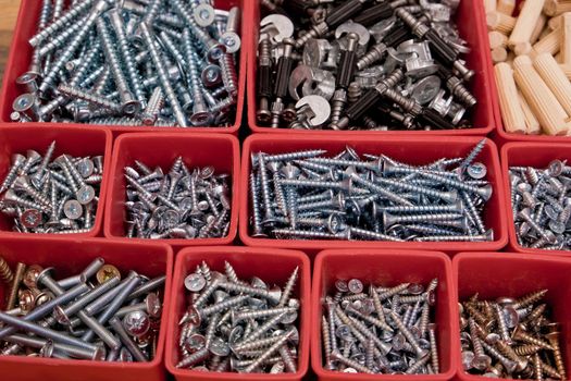 Various size and shape of screws, bolts, wooden pegs selection in red plastic tray box on a wooden background. Assembly kit for furniture. Selective focus.