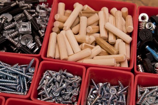 Various size and shape of screws, bolts, wooden pegs selection in red plastic tray box on a wooden background. Assembly kit for furniture. Selective focus.