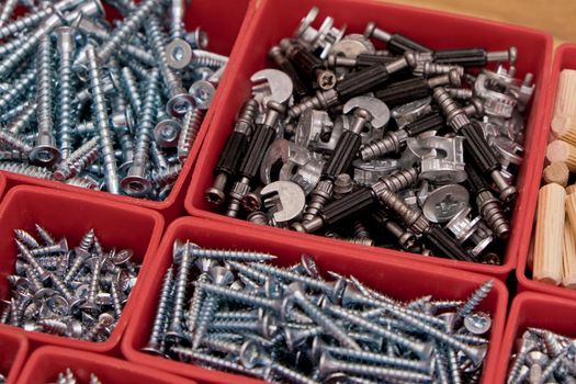 Various size and shape of screws, bolts, wooden pegs selection in red plastic tray box on a wooden background. Assembly kit for furniture. Selective focus.